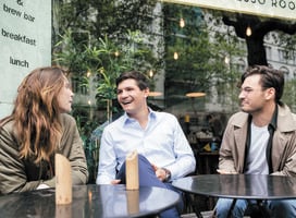 One female and two male postgraduate students sit at a table outside a cafe
