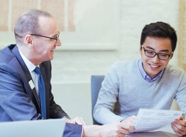 A male professor sits at a table next to a male student