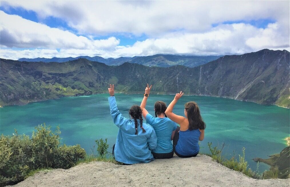 Three girls doing peace sign overlooking mountain and lake.