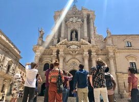 a group of students standing in front of Ortygia Cathedral, their backs are to the camera