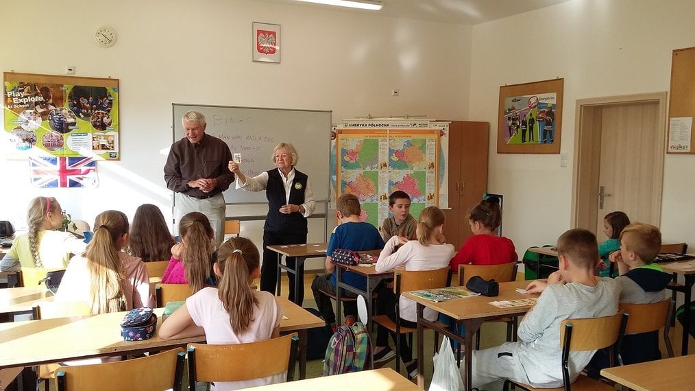 Two older adults stand at the front of a classroom of children.