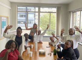 A group of people gathered around a table wave at the camera