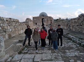 A group of 5 students stand in a row on an ancient paved road with ruins in the background.