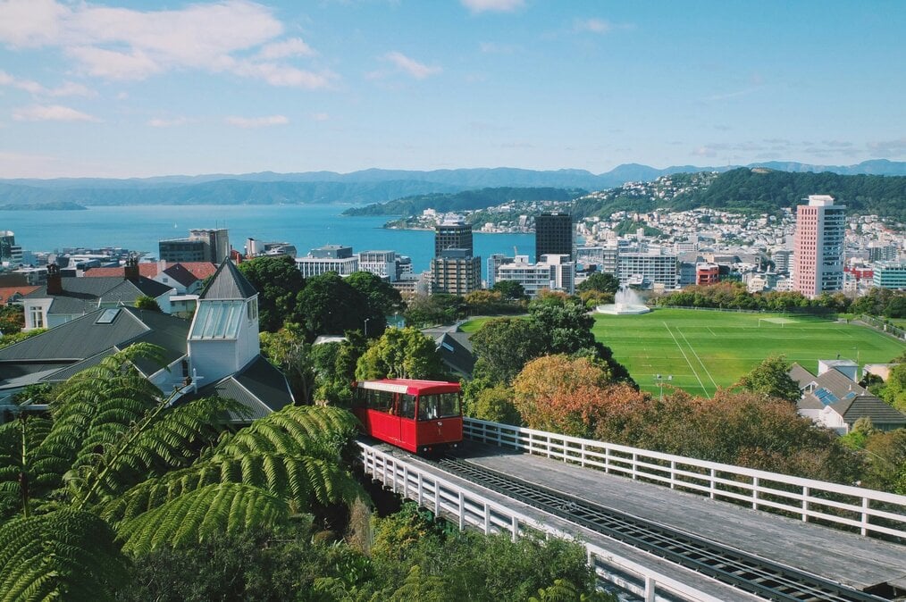 A red tram drives up a hill with a harbor in the background.