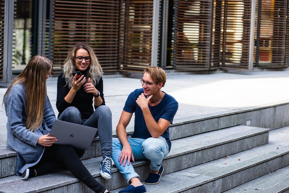 Three students sit on stairs outside with a laptop.