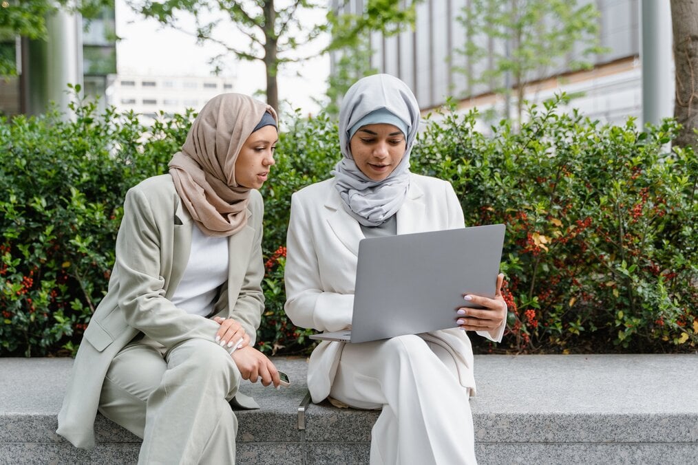 Two women with headscarves look at a laptop outside.