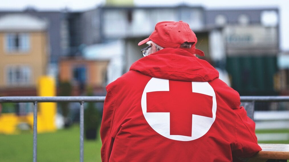 An older white man with his back to the camera wearing a red cross coat.