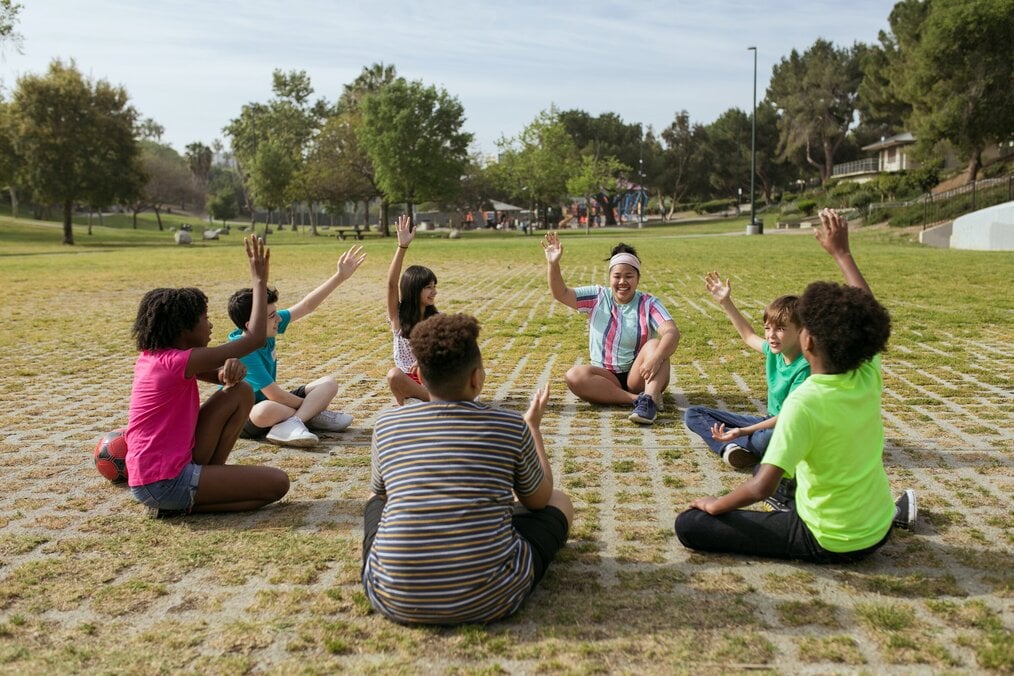 Teacher leading a class, sitting on ground with hand raised