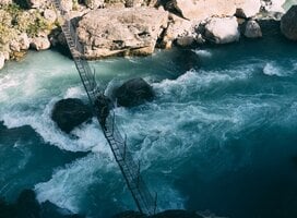 A NOLS student crosses a bridge over fast, blue water.