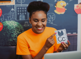 a woman in an orange shirt is smiling at a computer and pointing to a flashcard, which shows a picture of a cat and the letter C